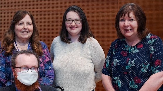 Three white adult women smiling and standing shoulder to shoulder plus one white man sitting in front wearing a medical face covering.