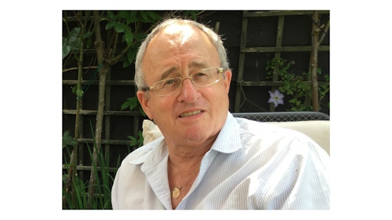 Steve smiles at the camera, he wears a white shirt and is wearing glasses. He is sat outside in the garden.