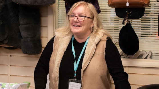 Volunteer Ruth, a lady with blonde hair and glasses, wearing a black top and cream gilet smiles at the shop counter at the Isabel Hospice shop