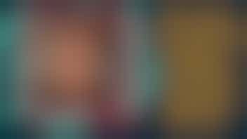 A selfie photo taken indoors showing someone wearing a cozy Fair Isle patterned sweater in browns, burgundy, and cream colors. They have long straight brown hair and are smiling warmly at the camera. In the background, there are decorative wall signs with text that includes phrases like "ALWAYS BE YOURSELF" and appears to have a unicorn theme. The lighting is warm and creates a homey atmosphere. The person appears to be in what looks like a home setting.