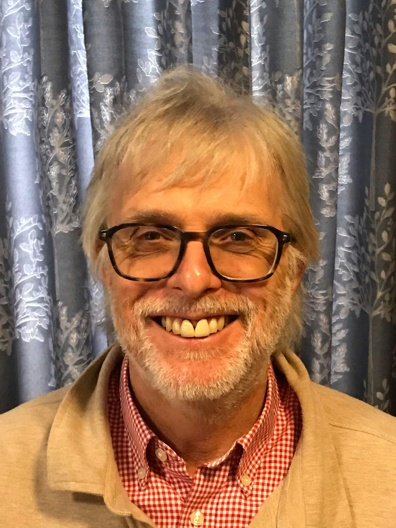 Nick has grey hair and is wearing black glasses, and a red checked shirt. He is smiling to the camera.