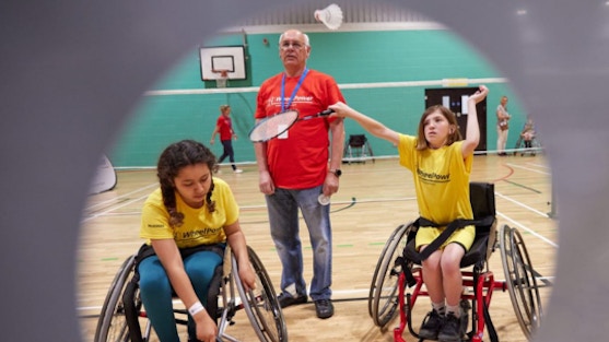 Man in red T shirt standing in between two children in sport wheelchairs who are playing badminton