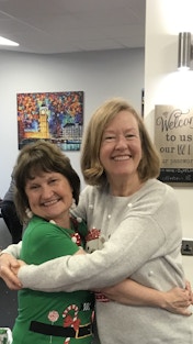 Image shows two volunteers dressed in Christmas jumpers hugging each other and smiling at the camera.