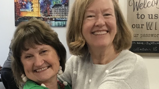 Image shows two volunteers dressed in Christmas jumpers hugging each other and smiling at the camera.